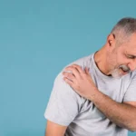 Stressed senior man with shoulder arthritis pain on blue backdrop